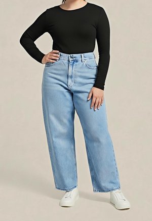 Femme portant un haut noir à manches longues, un jean taille haute bleu clair à jambes larges et des baskets blanches, debout avec une main sur la hanche.