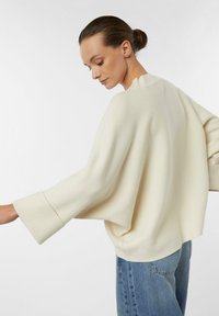Cream-colored oversized sweater with wide sleeves and a textured finish, paired with light blue jeans, showcasing a relaxed silhouette.