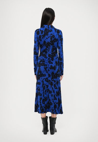 Uska haljina s dugim rukavima u plavoj boji s crnim apstraktnim uzorkom. Ima nabrani stražnji dio i suknju s faldicama. Uparena je s crnim čizmama.