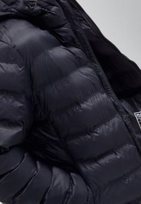 Chaqueta de plumas negra con costuras en forma de cuadrícula horizontal y cremallera visible parcialmente abierta que muestra el forro interior y una pequeña etiqueta de tela.