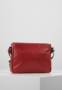 Červená obdélníková kožená krosbody taška s nastavitelným popruhem a stříbrným kováním, umístěná na bílé ploše na jednoduchém pozadí.