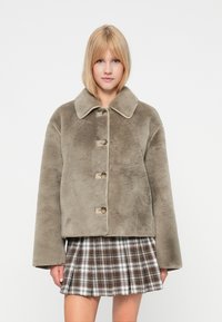 Veste cropped en fausse fourrure de couleur vert olive, avec un large col, cinq boutons et une texture douce, associée à une jupe à carreaux.