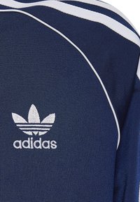 Tums zila sporta krekls izgatavots no teksturēta auduma, ar baltām svītrām un izšūtu baltu logo krūšu rajonā.