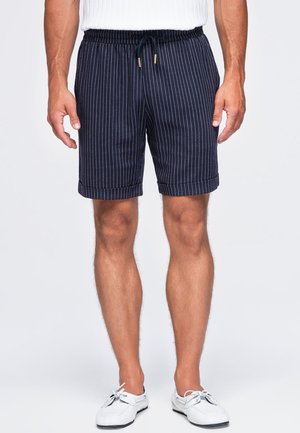 Mand iført marineblå knælange shorts med smalle striber og opkrogede kanter, kombineret med hvide bådsko og en hvid top, der er synlig ved taljen.