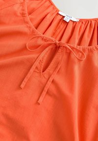 Jasnopomarańczowa bluzka z okrągłym dekoltem, z zapięciem na wstążkę oraz marszczonym wykończeniem przy dekolcie. Wykonana z lekkiej, fakturowanej tkaniny.