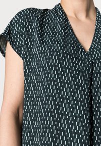 Tmavě zelený top s výstřihem do V, krátkými rukávy a texturovaným vzorem vertikálních světle zelených a černých pruhů. Hladká látka.