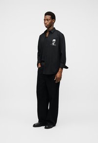 Νεαρός άνδρας που στέκεται, φορώντας ολόμαυρη άνετη ενδυμασία με γραφικό πορτρέτο στην τσέπη του πουκαμίσου, μπροστά σε απλό λευκό φόντο.