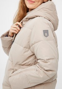 Lichtbeige pufferjack met een capuchon, voorzien van een ritszak en een logo-opdruk op de arm. Glad materiaal met een gestikte structuur.