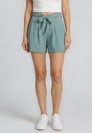 Frau trägt hoch taillierte salbeigrüne Shorts mit vorn gebundener Schleife, ein weißes Cropped-Top und weiße Sneakers, steht vor neutralem Hintergrund.