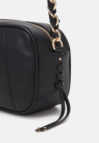 Borsa a tracolla in pelle nera con hardware dorato, caratterizzata da una tracolla intrecciata e chiusura con zip, superficie testurizzata e nodi decorativi laterali.