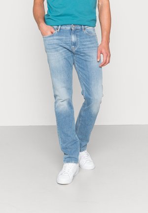 Mann trägt hellblaue ausgewaschene Jeans, türkisfarbenes Hemd und weiße Turnschuhe, steht mit einer Hand in der Tasche vor einfarbigem Hintergrund.