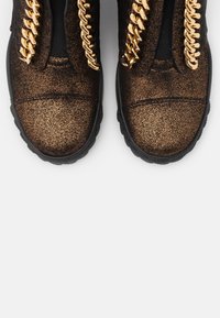 Paire de chaussures brun foncé scintillantes avec des semelles noires épaisses et de grands lacets en chaîne dorée brillante sur fond blanc.
