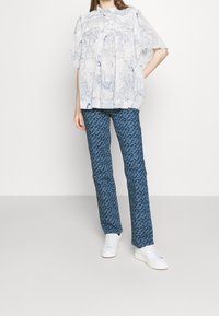 Blusa bianca a fantasia con motivo floreale blu e maniche arricciate. Pantaloni blu a fantasia con una forma aderente. Indossati con sneaker bianche.