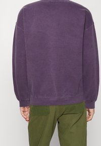 Lila sweatshirt med långa ärmar, ribbade manschetter och nederkant, med en avslappnad passform. Bärs med gröna byxor som har bakfickor.