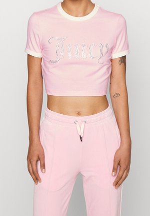 Osoba ubrana w jasnoróżowy krótki top z napisem „Juicy” z kryształków oraz pasujące jasnoróżowe spodnie dresowe ze ściągaczem, odsłaniająca brzuch i tatuaże na ramionach.