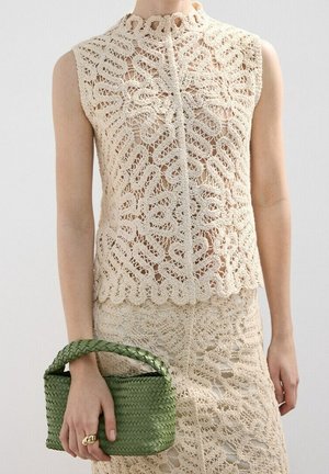 Ensemble top sans manches beige en crochet et jupe porté, tenant un petit sac à main vert tressé avec une courte poignée dans la main gauche.