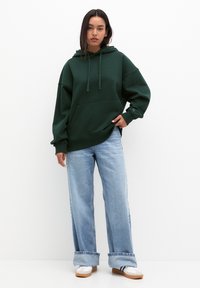 Dunkelgrüner oversized Hoodie mit einer Fronttasche und Kordelzug. Kombiniert mit hellblauen, weit geschnittenen Jeans und weißen Turnschuhen.