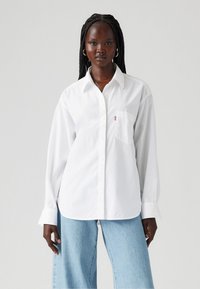 Biała koszula zapinana na guziki z bawełny, z kieszonką na piersi, kołnierzykiem i długimi rękawami. Połączona z jasnoniebieskimi spodniami dzwonami.