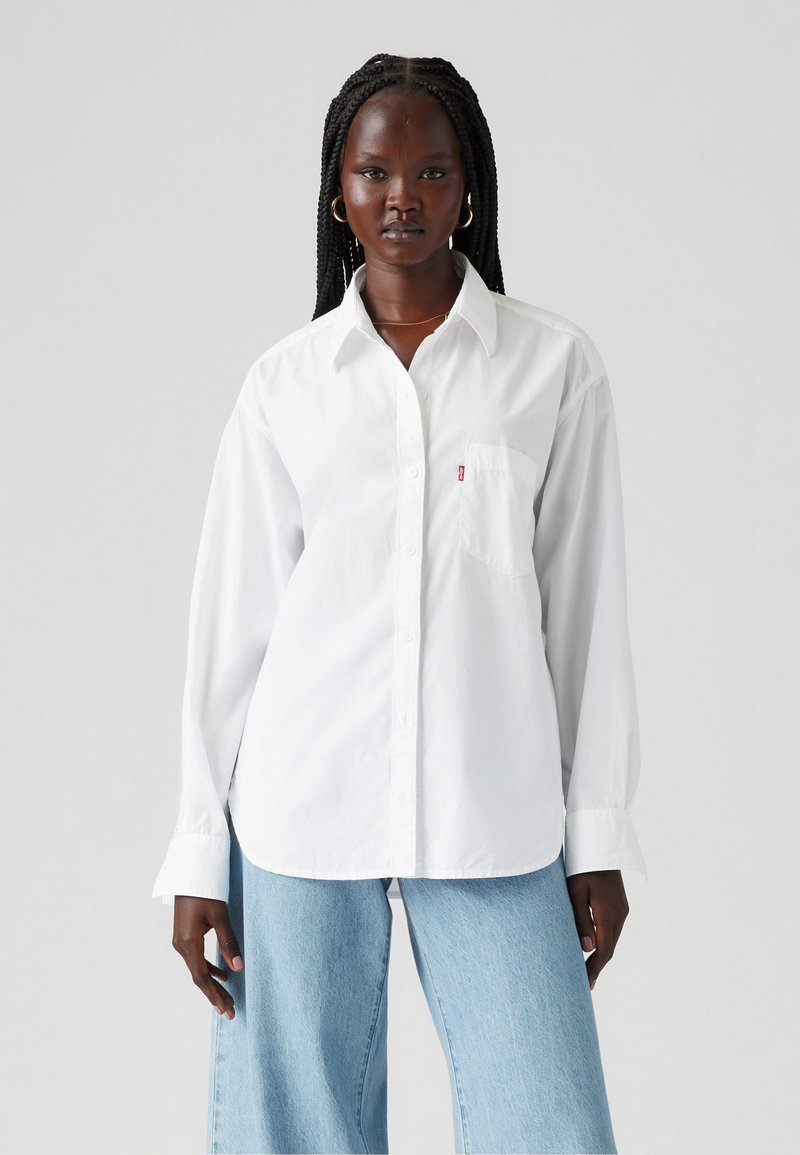 Biała koszula zapinana na guziki z bawełny, z kieszonką na piersi, kołnierzykiem i długimi rękawami. Połączona z jasnoniebieskimi spodniami dzwonami.