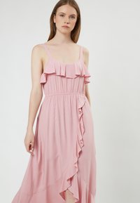 Vestido sin mangas de color rosa con escote fruncido y detalle de volantes en el costado. Hecho de una tela ligera, cuenta con una cintura elástica para mayor comodidad.