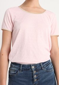 Camiseta corta de manga corta en rosa claro con un patrón texturizado y un ribete dorado alrededor del escote, combinada con jeans de mezclilla oscuros con botones.