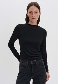 Frau mit schulterlangen, dunklen Haaren trägt ein enges schwarzes langärmeliges Oberteil und hochgeschnittene schwarze Jeans vor einem einfarbigen Hintergrund.