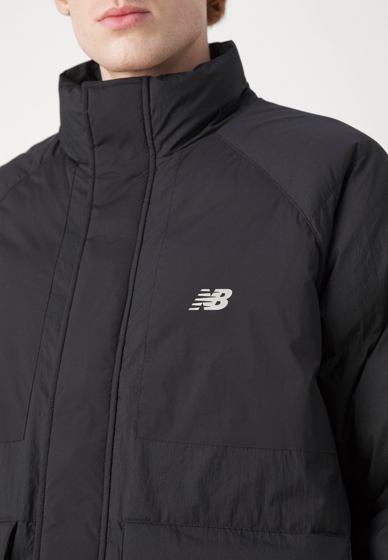 Mann trägt eine schwarze New Balance gesteppte Jacke mit hohem Kragen vor einem weißen Hintergrund.