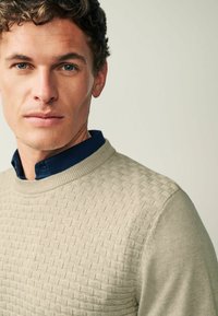 Pull en beige texturé avec un motif tissé, doté d'un col rond et d'une chemise col bleu marine en dessous. Finition en tissu lisse.