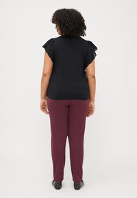 A person with curly hair wearing a black top and maroon pants stands with their back facing the camera, showcasing a simple outfit.