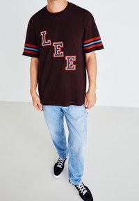 Camiseta marrón con mangas a rayas rojas y azules, con grandes letras blancas "LEE" al frente. Combinada con vaqueros azul claro.