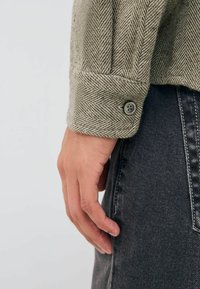 Herringbone-patterned shirt in light brown with a buttoned cuff, paired with dark denim jeans. The fabric appears textured and thick.