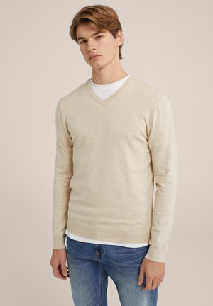 Beige V-Ausschnitt-Pullover aus weichem Material, mit langen Ärmeln und tailliertem Design, über einem weißen T-Shirt getragen, kombiniert mit blauen Jeans.