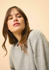 Pull gris en tissu doux et texturé, avec un col rond et des épaules tombantes, porté par une personne aux longs cheveux bruns sur un fond clair.