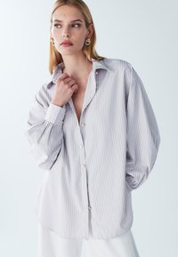 Helles, gestreiftes, locker geschnittenes Button-up-Hemd mit Kragen, goldenen Knöpfen und voluminösen Ärmeln. Glatte Stoffstruktur.