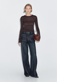 Dark brown fitted long-sleeve top paired with high-waisted, wide-leg dark denim jeans featuring contrasting stitching and back pockets. Holding a small brown bag.