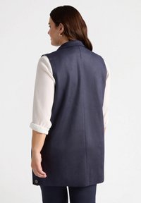 Marineblauer ärmelloser Anzug mit glatter Textur, tailliertem Design und seitlichen Knopfakzenten, kombiniert mit einem cremefarbenen Langarmshirt.