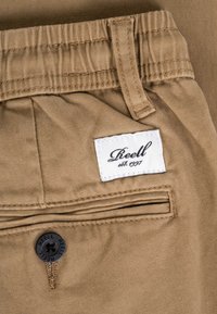 Primer plano del bolsillo de unos pantalones marrones con un botón negro etiquetado "Reell" y una etiqueta de tela blanca de la marca que dice "Reell est. 1997."
