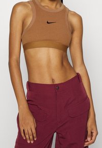 Brun ribbad sportbehå med en svart Nike-logo, med en racerback-design och en bred, texturerad underband, parat med djup röda cargo-byxor.