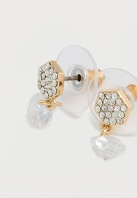 Επεξεργασμένα με χρυσή απόχρωση εξαγωνικά σκουλαρίκια με διάφανα στρας στην κορυφή, διακοσμημένα με κρεμαστά διαφανή κρυσταλλικά στοιχεία. Περιλαμβάνονται διαφανείς πίσω στήλες.