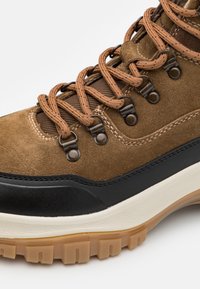 Brązowy zamszowy but z syntetycznymi czarnymi akcentami, wyraźnym systemem sznurowania, gumową podeszwą i teksturowanym wykończeniem na śródpodeszwie.