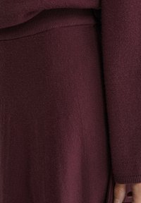 Bordeauxfarbener Strickstoff mit strukturierter Oberfläche; verfügt über einen breiten Bund und eine leicht ausgestellte Silhouette, die weiches, warmes Material präsentiert.
