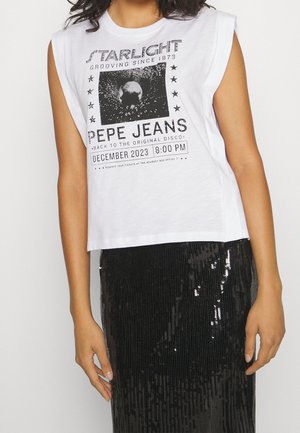 Femme portant un haut blanc sans manches avec un texte imprimé noir et une jupe noire à sequins, debout devant un fond uni.