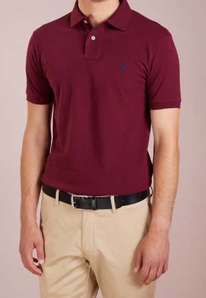 Bordeauxrotes Poloshirt mit kurzen Ärmeln, zweiknöpfigem Schlitz und blauem Logo. Kombiniert mit khakifarbenen Hosen und einem schwarzen Gürtel. Glatte Textur.