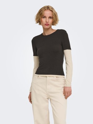 Femme aux cheveux blonds courts portant un haut noir à manches courtes superposé à une chemise beige à manches longues et un pantalon beige, debout devant un fond blanc.