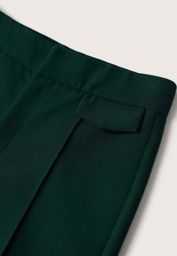 Détail d’un pantalon en tissu vert foncé montrant la taille et le petit rabat de poche sur un fond clair.