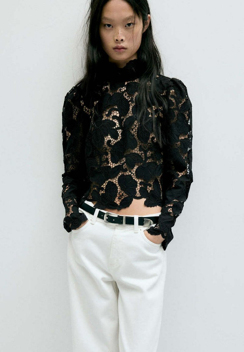 Femme aux longs cheveux foncés portant un haut noir en dentelle florale à manches longues et un pantalon blanc avec une ceinture noire, mains dans les poches, fond neutre.