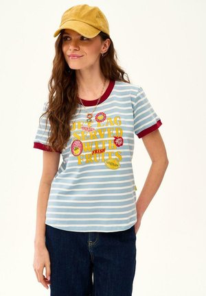 Kobieta nosząca żółtą czapkę, niebiesko-biały pasiasty t-shirt z czerwonym obszyciem i tekstem o tematyce owocowej, w połączeniu z ciemnoniebieskimi jeansami.