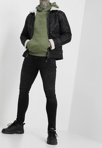 Giubbotto di jeans nero con colletto in shearling bianco, felpa verde sotto e jeans skinny neri con texture. Sneakers sportive nere.