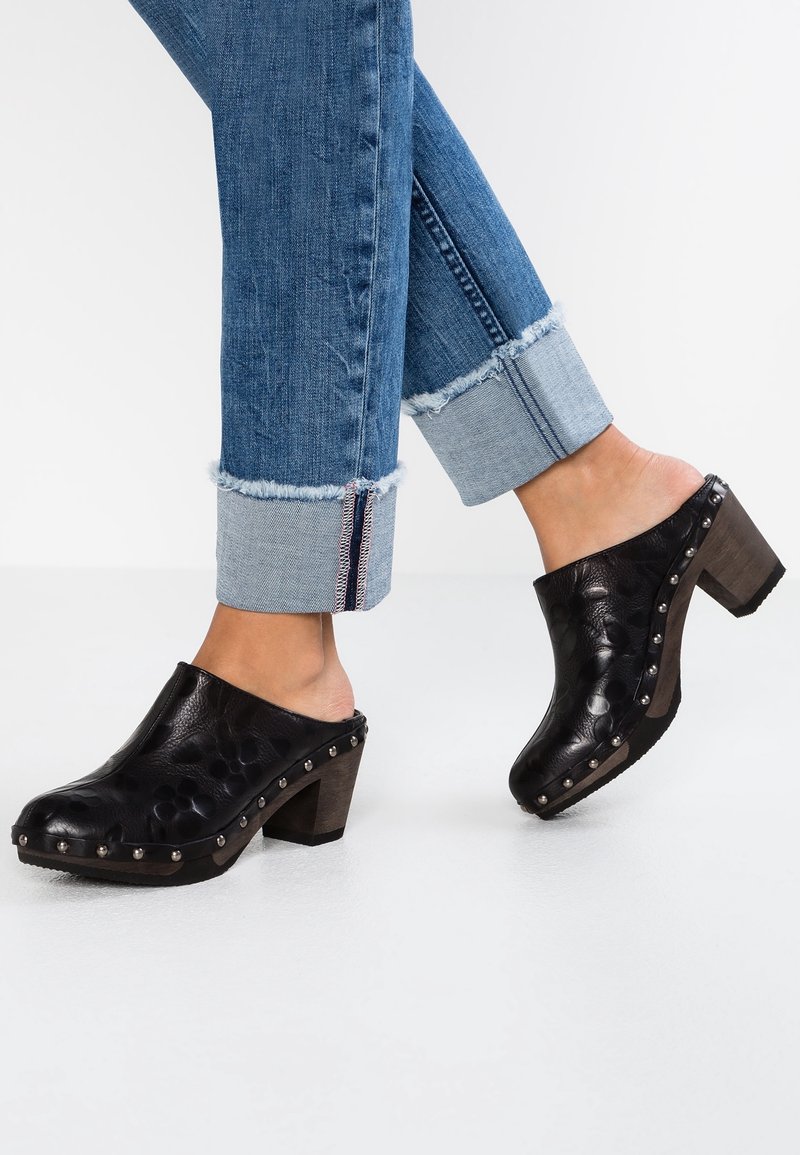 Personne portant un jean bleu retroussé et des sabots à talons en cuir noir avec des détails cloutés sur fond blanc.