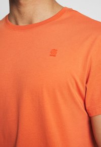 Camiseta de algodón naranja con cuello redondo, que presenta un pequeño logo bordado en el pecho. La tela tiene una textura suave y ligera.